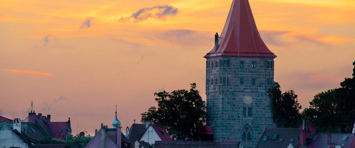 A tower with a red roof and a clock, surrounded by houses with red roofs, against a sunset sky.