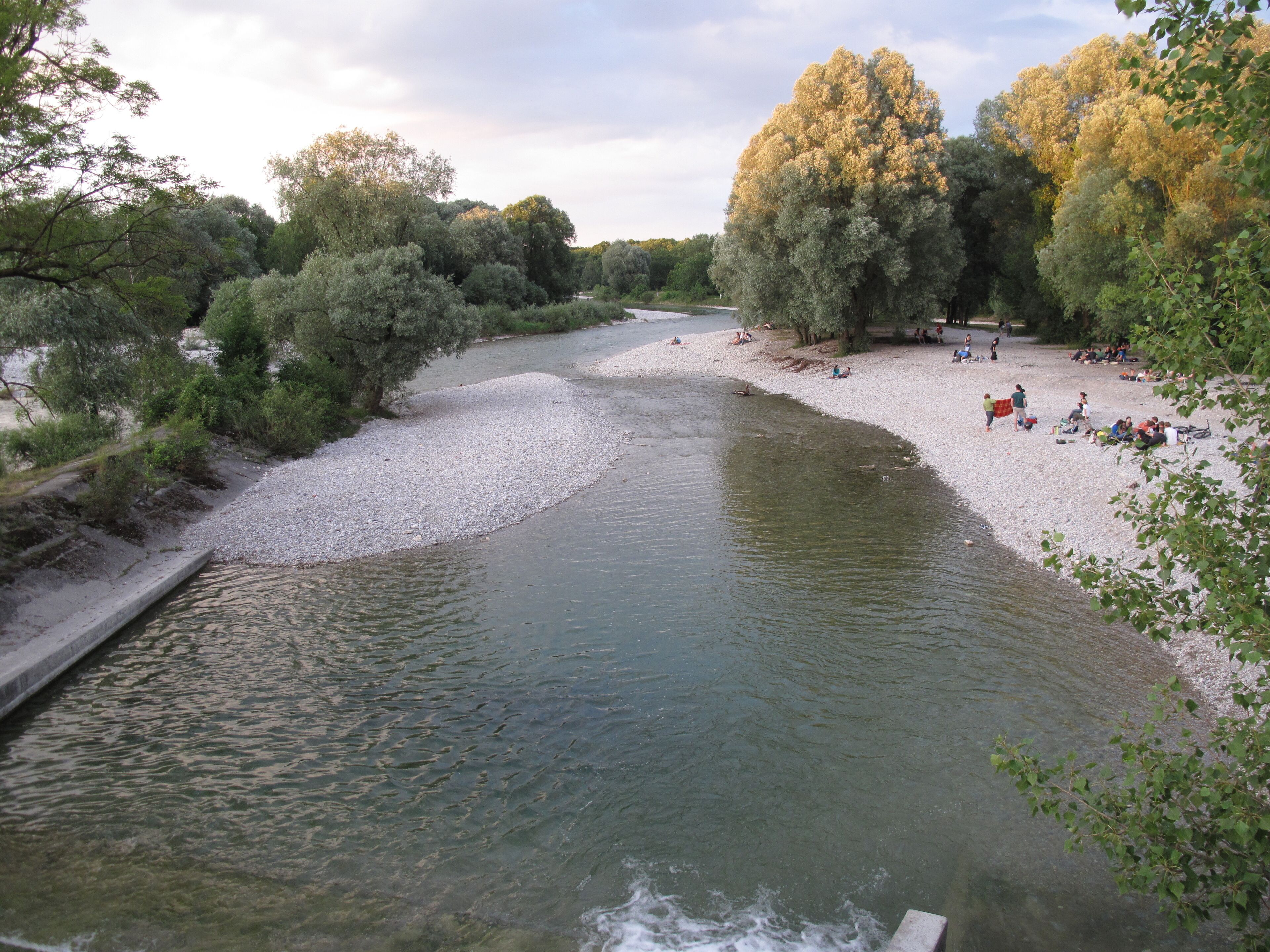 A river with a pebbly bank, surrounded by trees and a sandy beach with people relaxing.