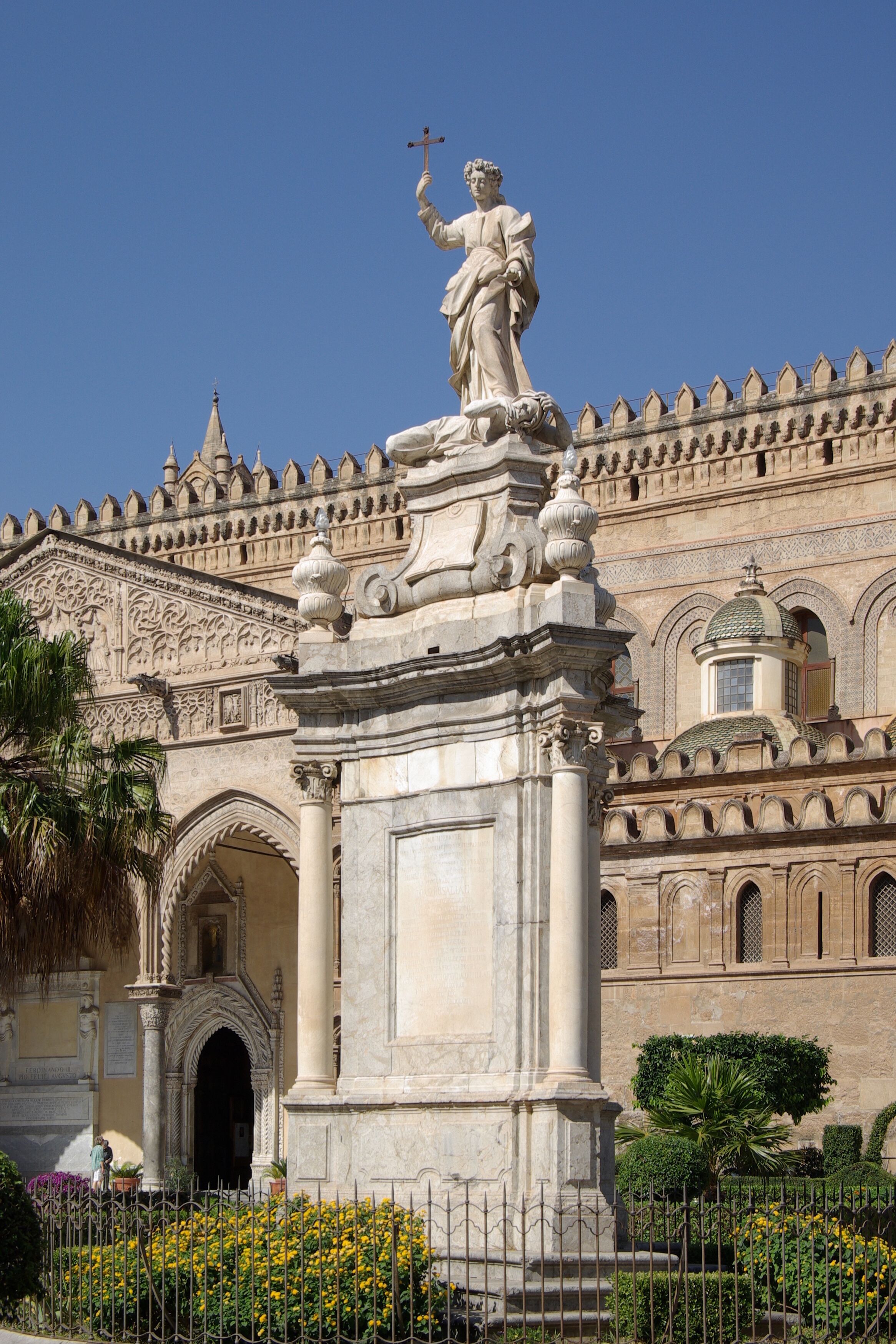 Une statue représentant une femme tenant une croix, une fontaine et un bâtiment à l’architecture complexe.