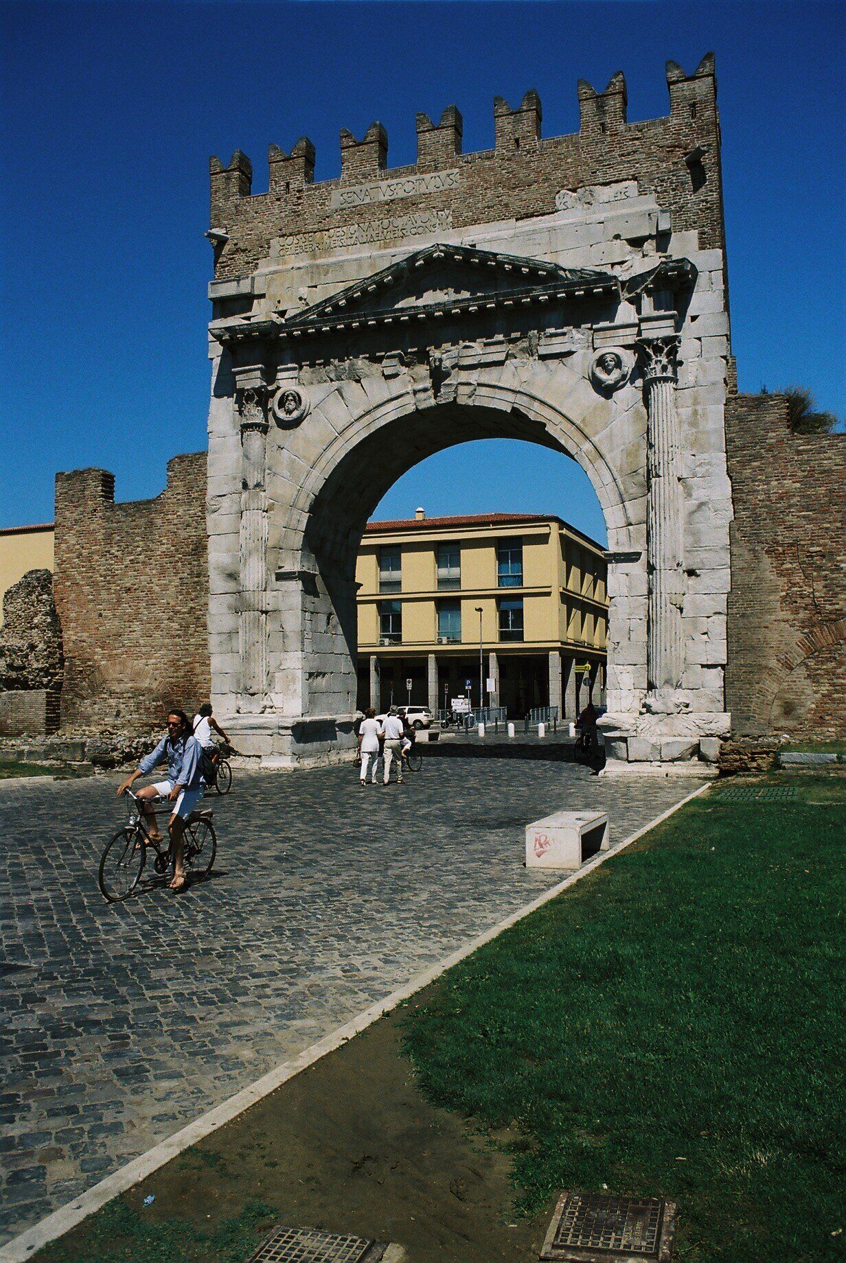 An ancient stone archway with inscriptions, a cobblestone street, and a building in the background.