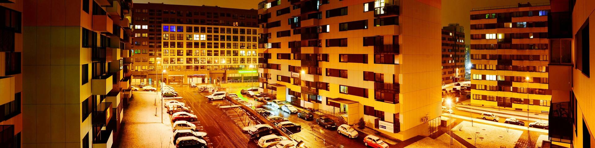 A nighttime urban street with multi-story buildings and parked cars.