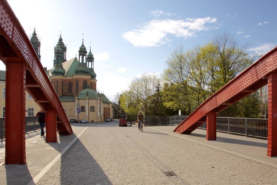 Uma ponte para pedestres com estruturas de metal vermelhas, um caminho de pedra e uma igreja com cúpulas verdes ao fundo.