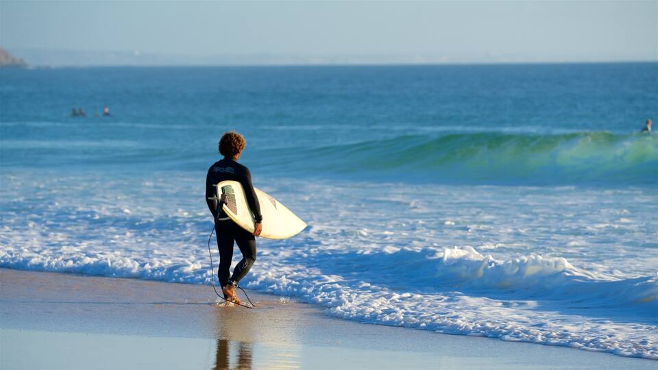 A surfer walking on the beach with a surfboard, waves in the background, and a clear sky.