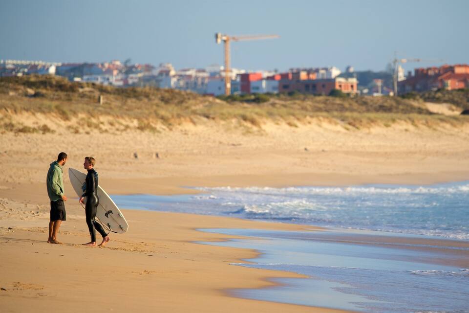 Zwei Personen laufen auf einem sandigen Strand, im Hintergrund sind Wellen, ein Windrad und Gebäude zu sehen.