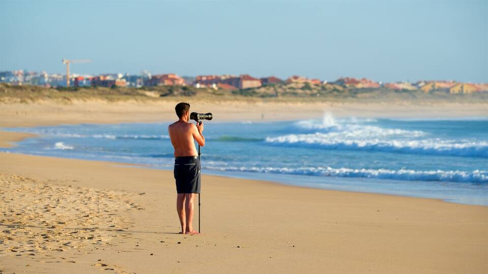 A man taking a photo on a sandy beach with waves in the background and buildings in the distance.
