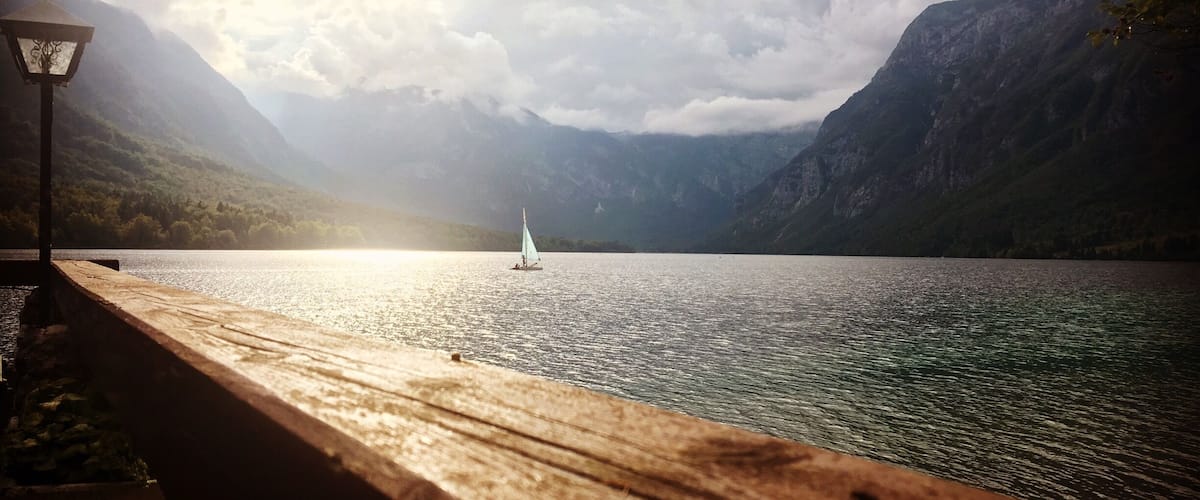A wooden dock extending into a calm lake with a sailboat in the distance, surrounded by mountains and a partly cloudy sky.
