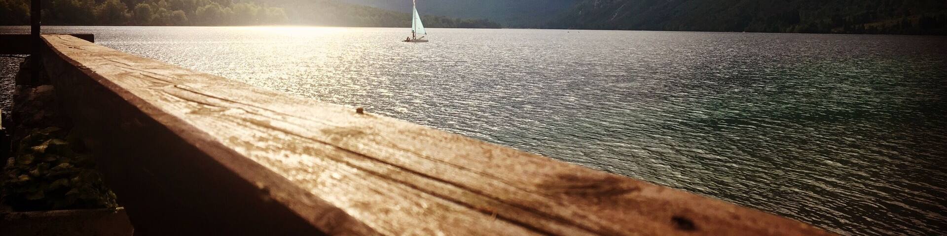 A wooden dock extending into a calm lake with a sailboat in the distance, surrounded by mountains and a partly cloudy sky.