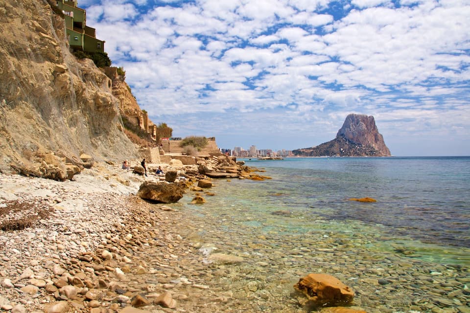 A rocky beach with clear water, a cliff, and a distant mountain.