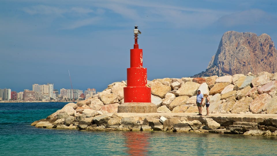 A red lighthouse on a rocky shore with a city skyline and a mountain in the background.