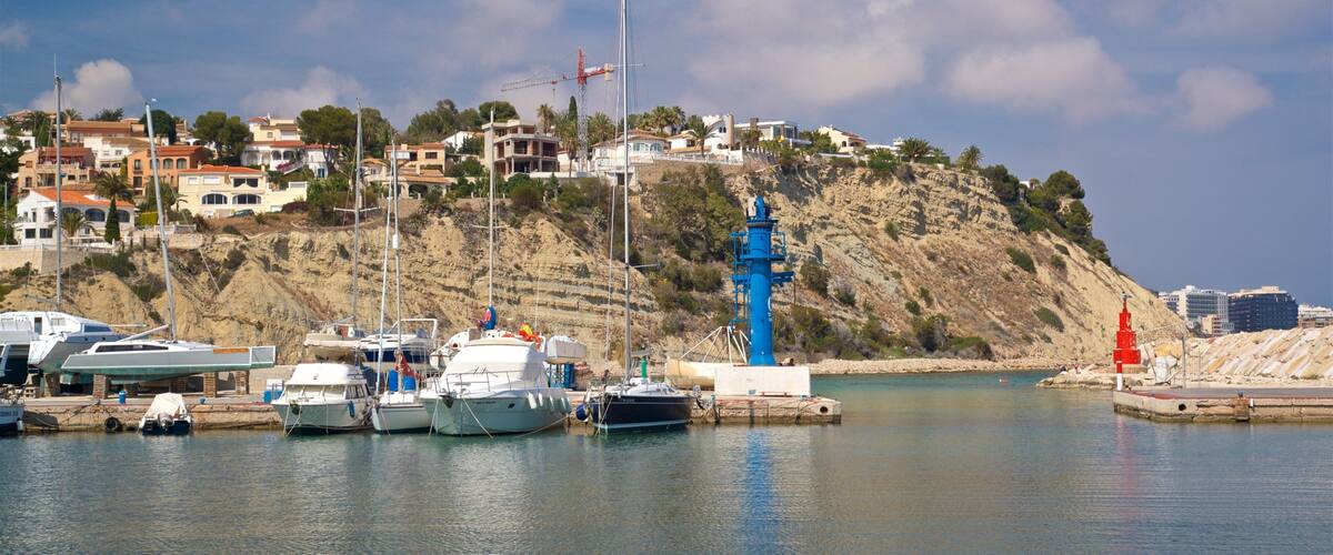 A coastal marina with boats docked, a prominent blue lighthouse, and a hillside with buildings.