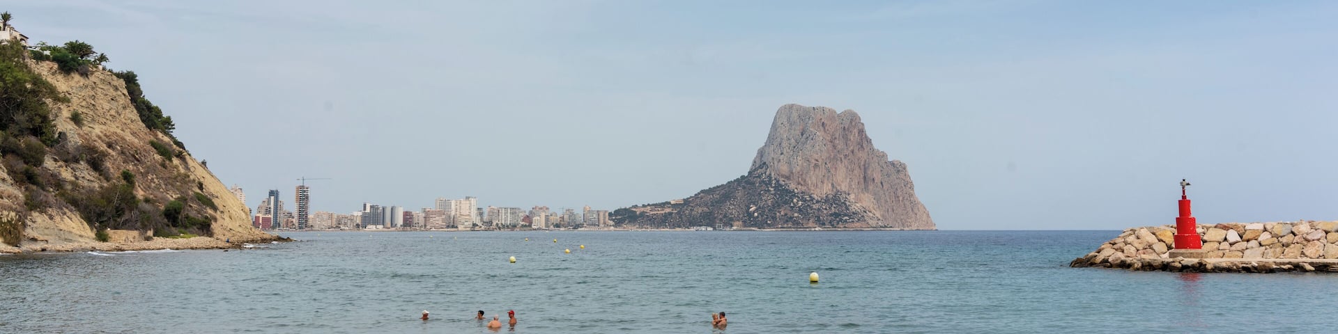 A coastal scene with people swimming, a rocky outcrop, and a city skyline in the background.