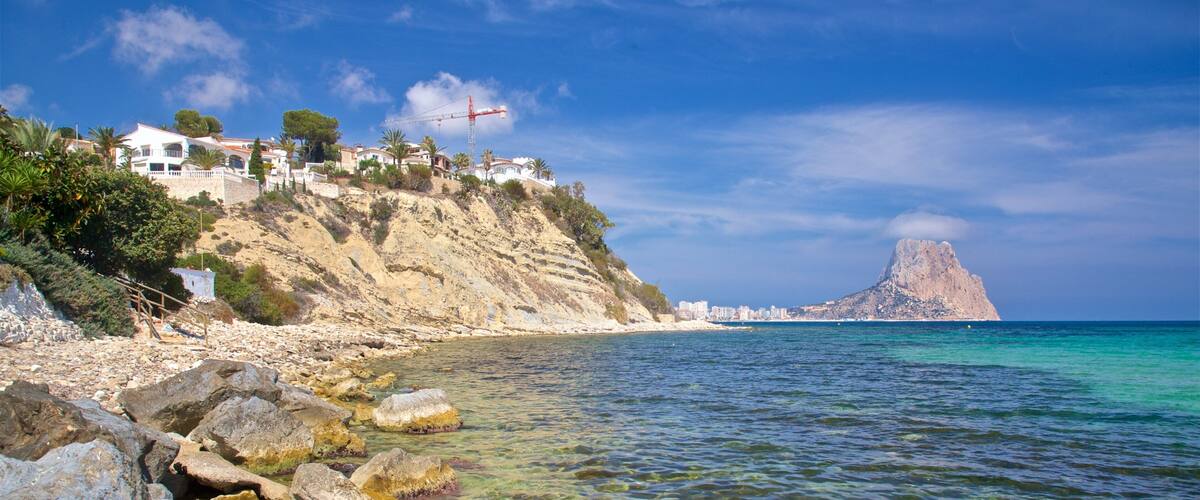 A coastal landscape with clear waters, rocky shore, and a prominent mountain peak in the distance.