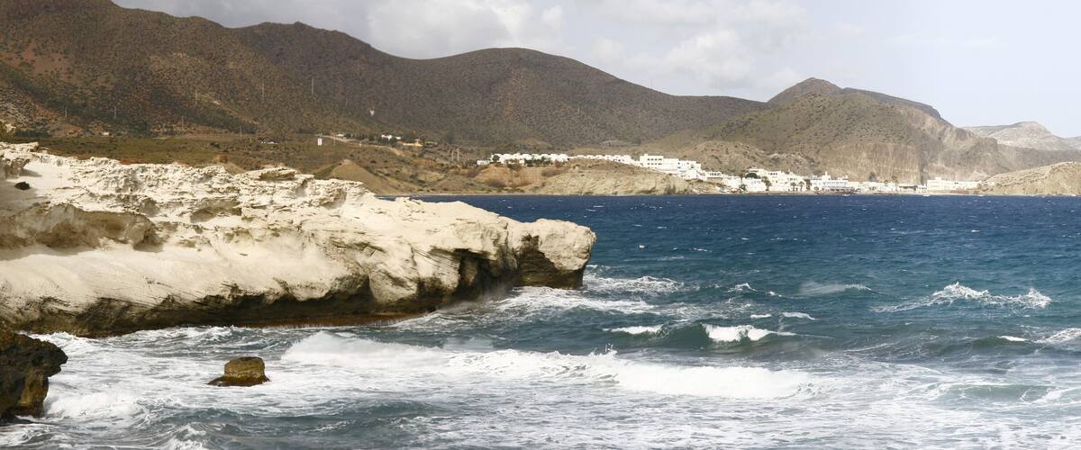 A coastal landscape with white buildings, rocky cliffs, and a mountainous backdrop.