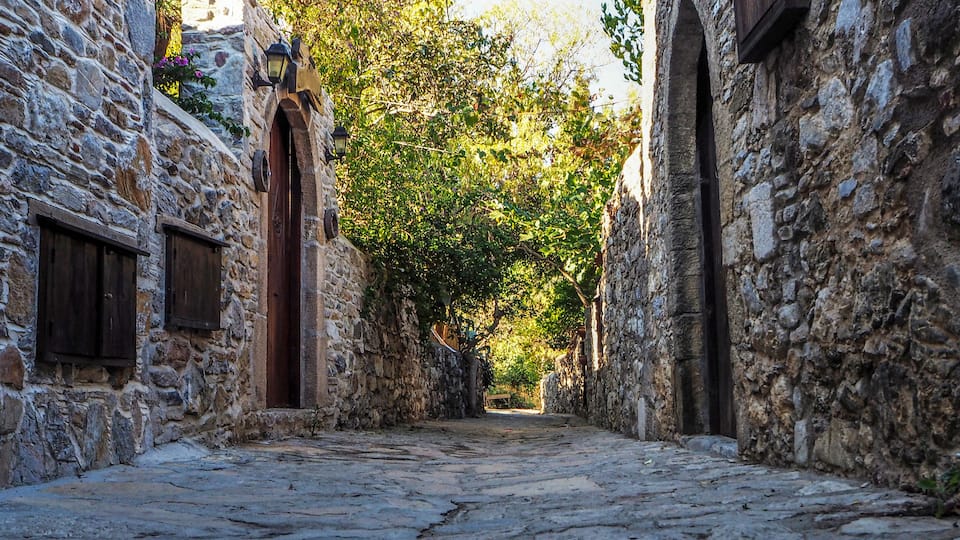 A narrow stone-paved alley flanked by rustic stone buildings with wooden shutters and a wooden door.