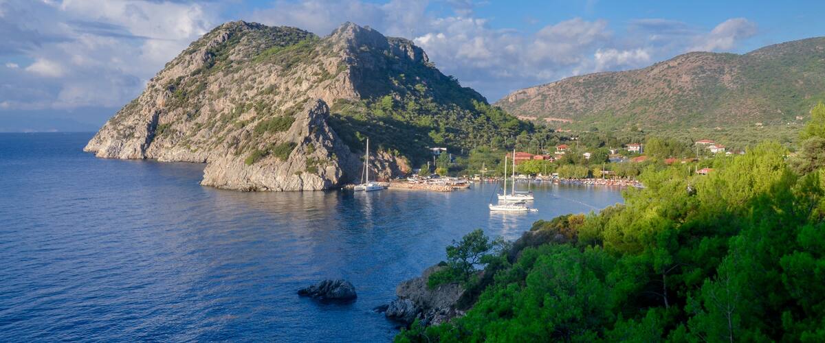 A coastal landscape with a prominent rocky island, a marina with boats, and a village with red-roofed houses.