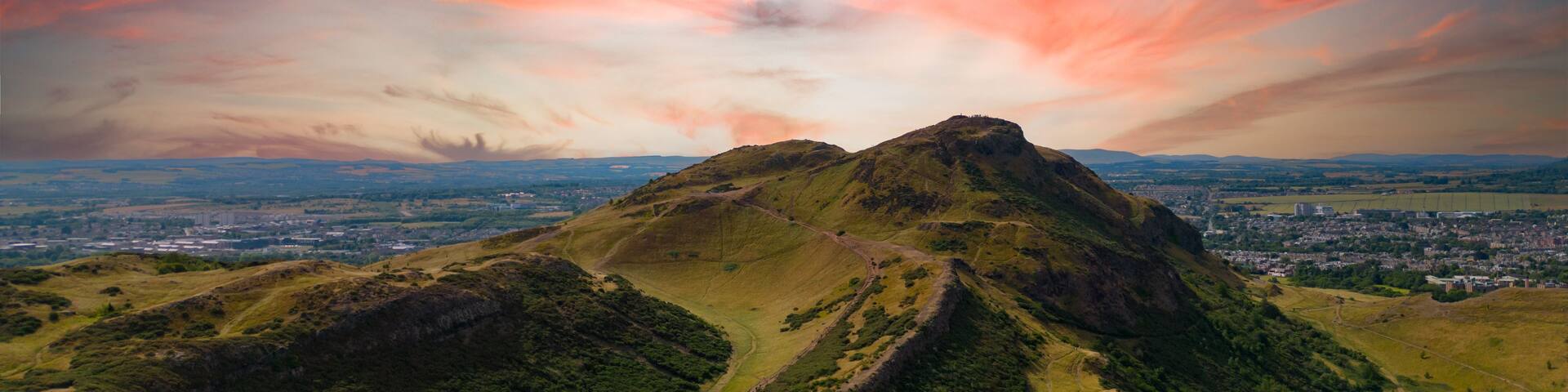 A mountain with a clear path leading to its peak, surrounded by lush greenery and a distant cityscape under a colorful sky.