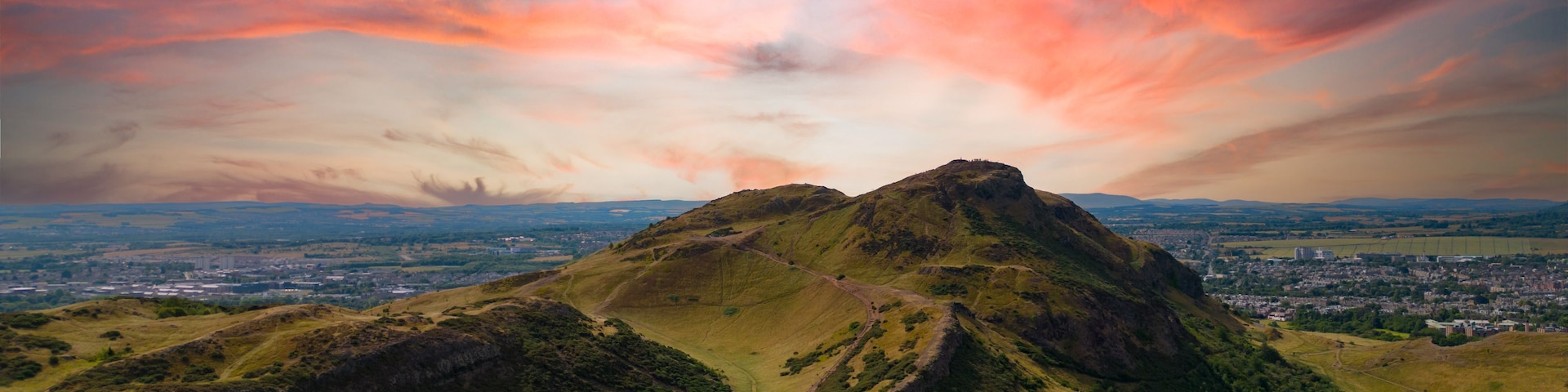 Montanha com trilha bem definida até o cume, cercada por vegetação exuberante e uma paisagem urbana distante sob um céu colorido.