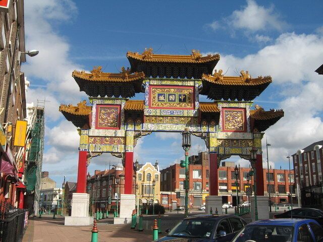 A traditional Chinese gate with ornate decorations and red pillars, set in an urban street with buildings and cars.