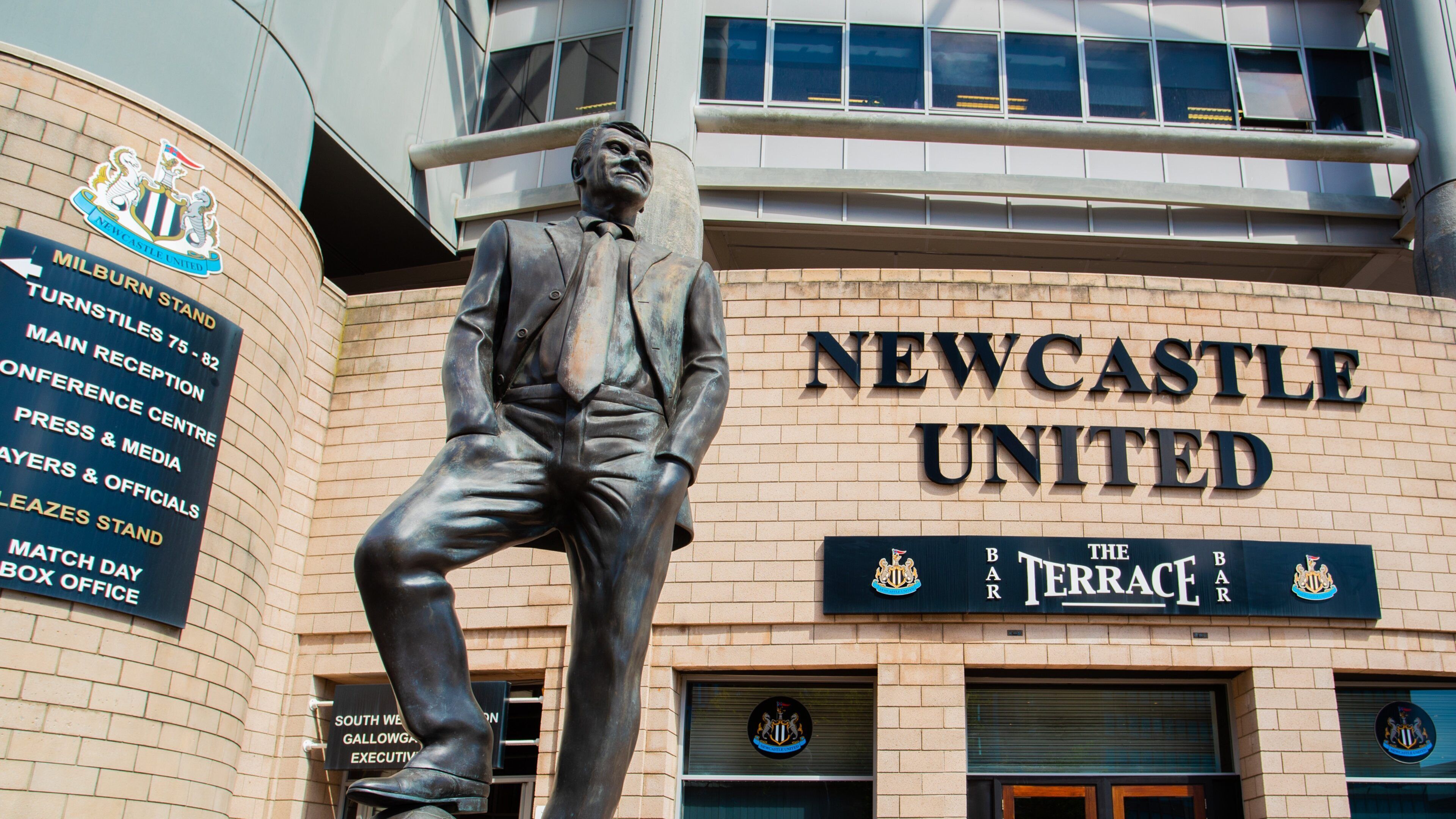 En statue af en mand i jakkesæt og slips, et skilt for Newcastle United, og terrassen ved The Bank of England.