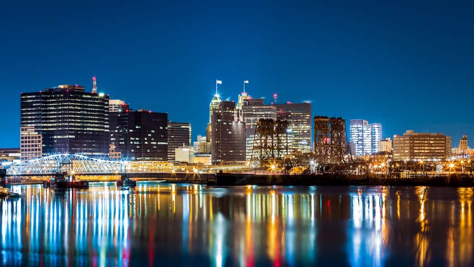 A city skyline at night with illuminated buildings and a bridge reflecting on the water.