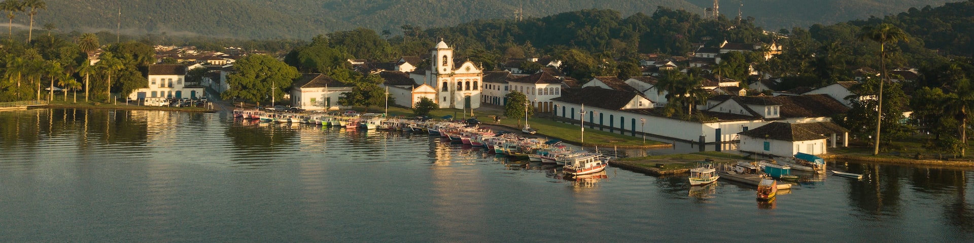 A coastal town with a prominent church, boats docked along the waterfront, and a backdrop of mountains.