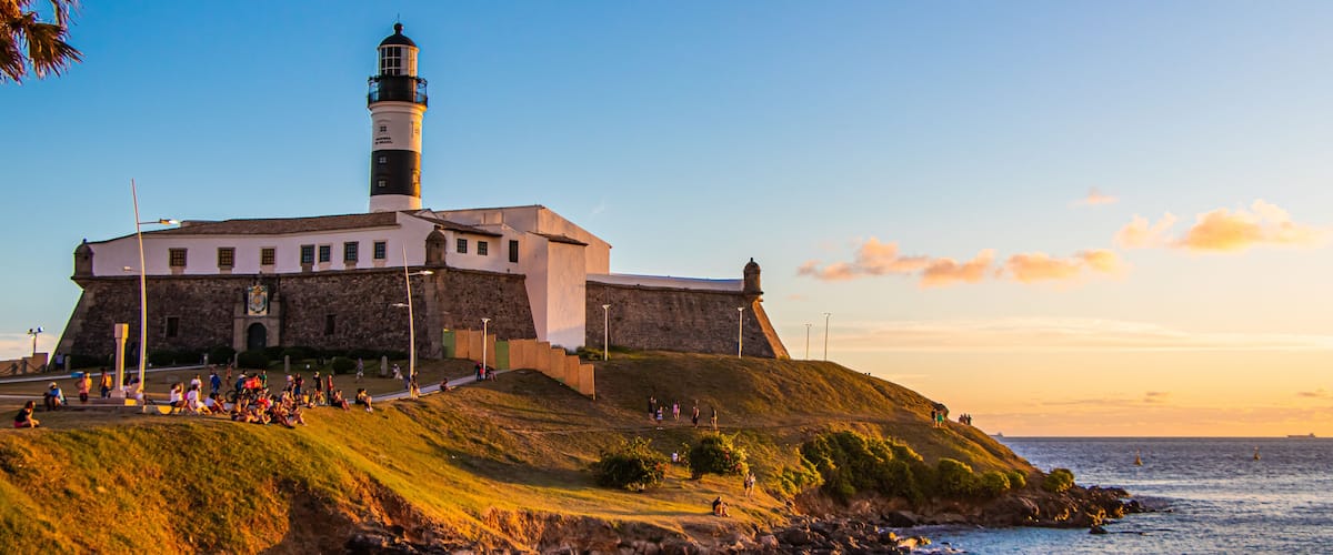 A lighthouse on a grassy hill overlooking the sea, with people walking around and a clear sky.