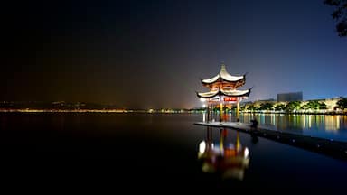 Un pabellón chino tradicional junto a un lago tranquilo, reflejando la estructura y las luces de la ciudad.