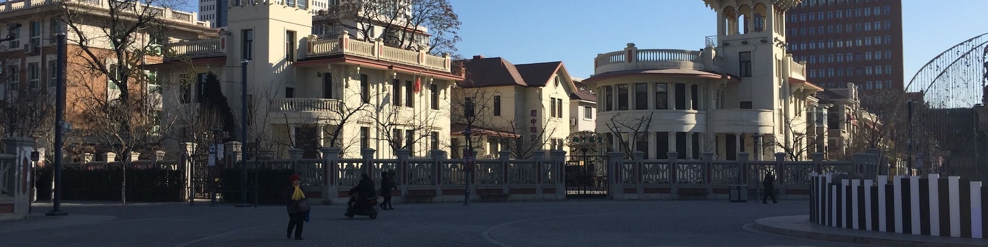 Uma praça de pedra com edifícios históricos, incluindo uma torre imponente e uma estrutura clássica com colunas.
