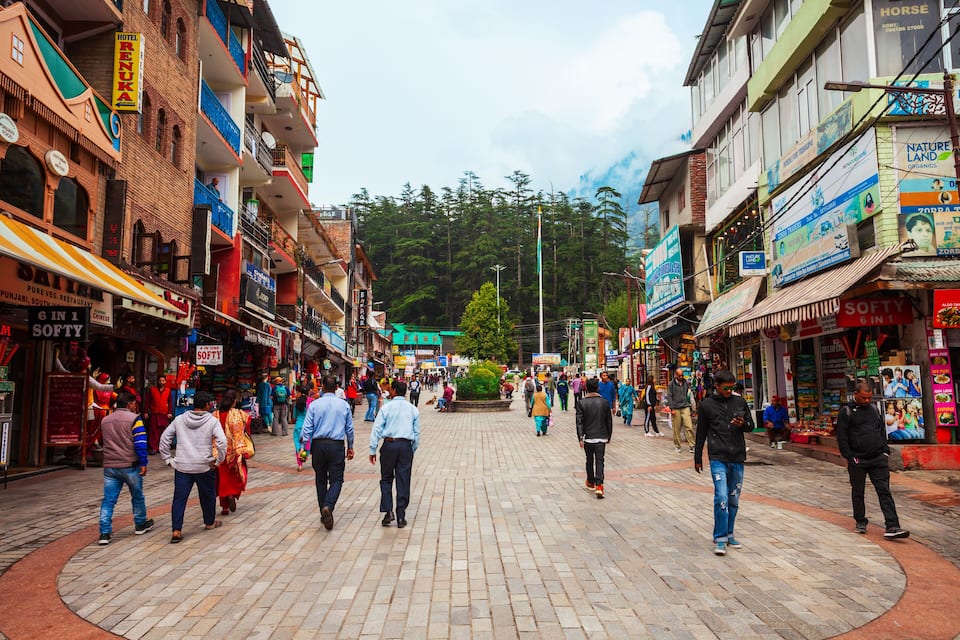 Una calle animada con tiendas y restaurantes, una fuente central y montañas al fondo.