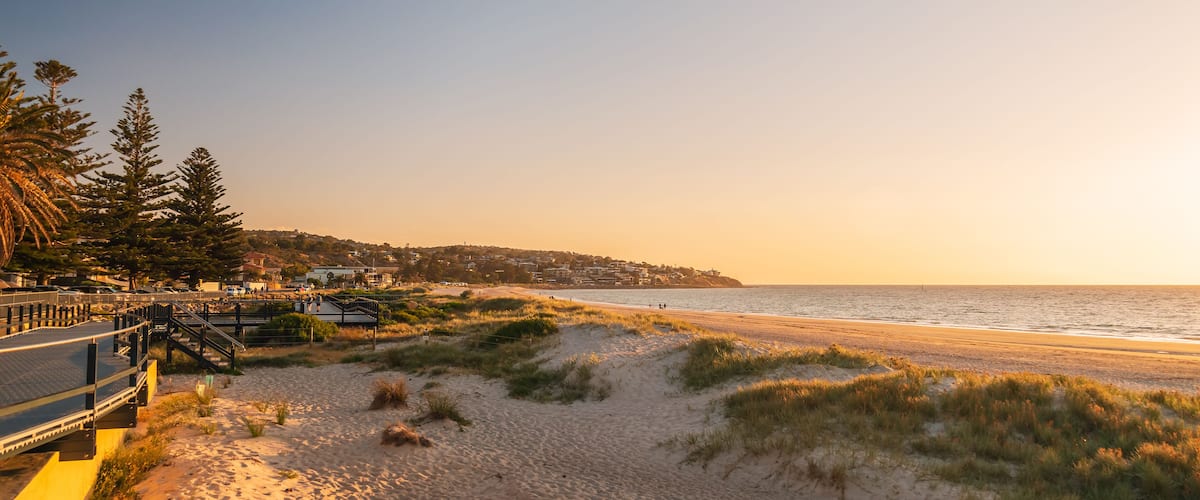 Una platja amb dunes de sorra, una passera de fusta i un poble costaner al fons.