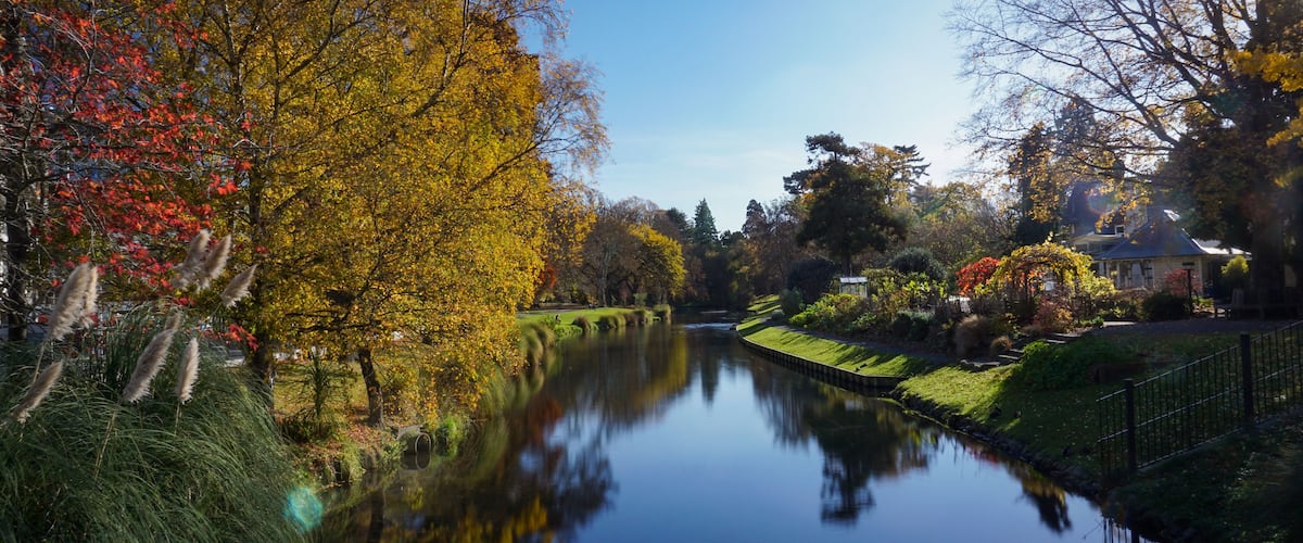 Une rivière paisible où se reflètent les arbres aux couleurs automnales, une rive herbeuse bien entretenue et une maison avec un jardin.