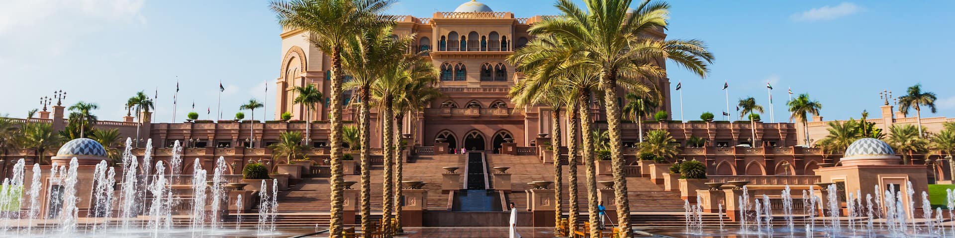A grand building with a fountain in front, palm trees, and a clear blue sky.