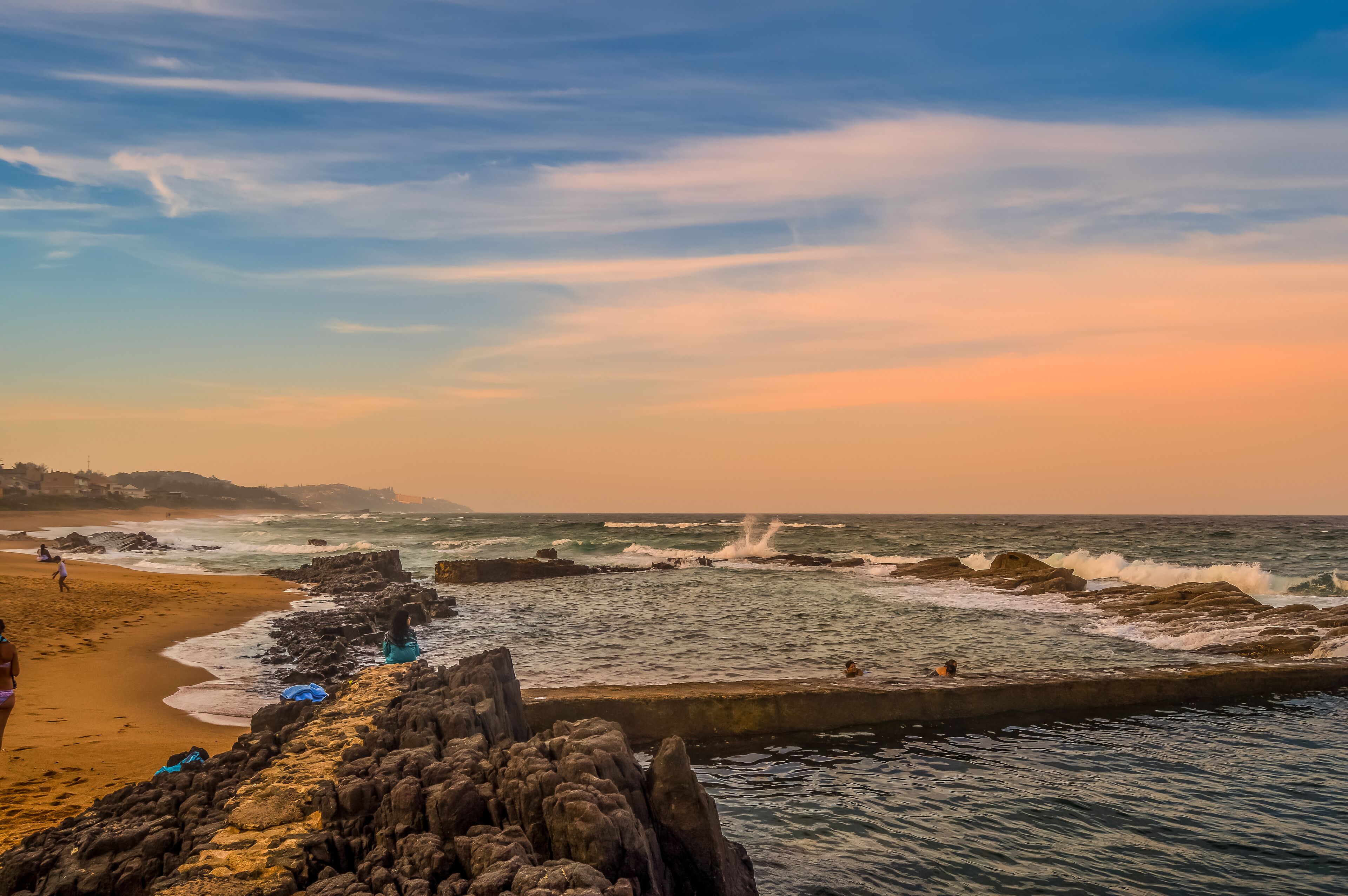 A rocky coastline with waves crashing against the shore, a sandy beach, and a clear sky.