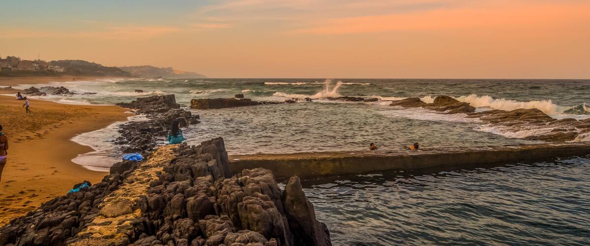 A rocky coastline with waves crashing against the shore, a sandy beach, and a clear sky.
