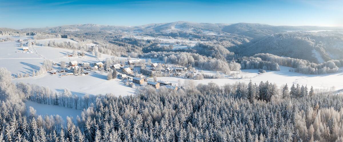 A snow-covered village nestled in a valley with dense forests and distant mountains.