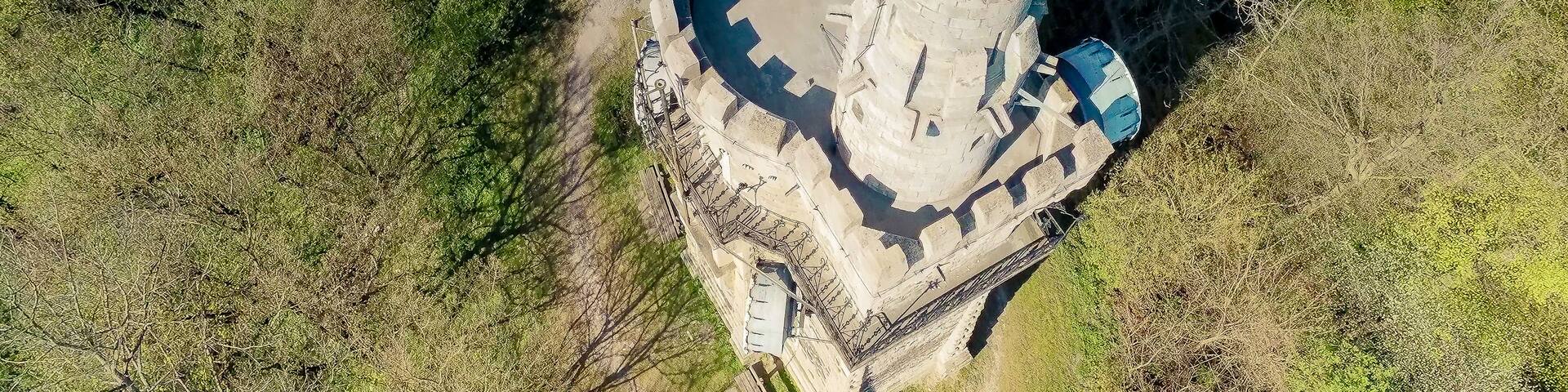 An aerial view of a historical tower with a circular base and multiple domes, surrounded by dense greenery.