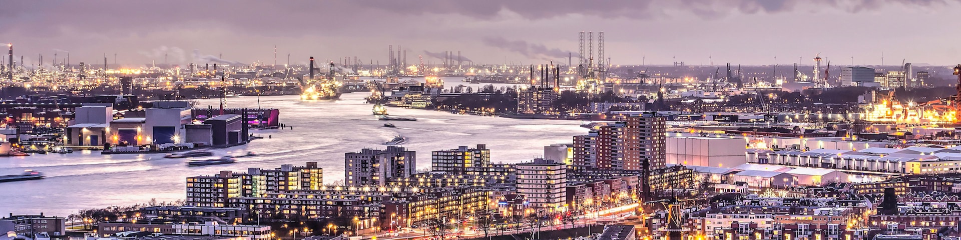 Paisagem urbana ao entardecer, com um rio, edifícios industriais e uma ponte.