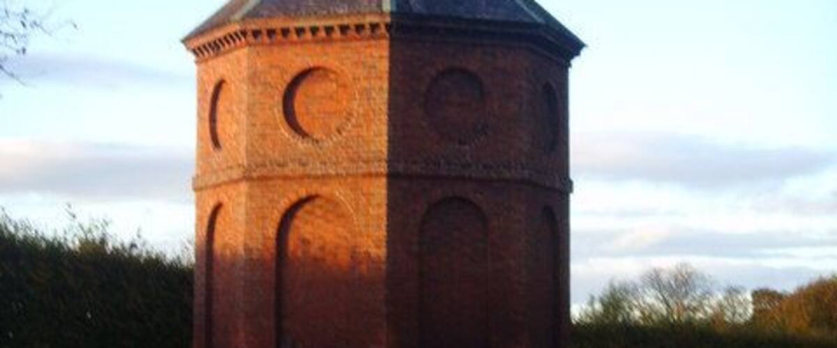 A brick tower with a conical roof and circular windows, surrounded by a grassy field.