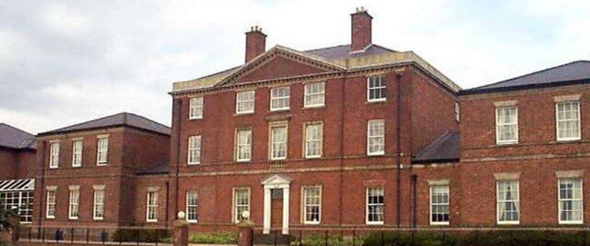 A large brick building with a symmetrical facade, a central entrance, and a well-manicured lawn with a gravel path leading to the entrance.