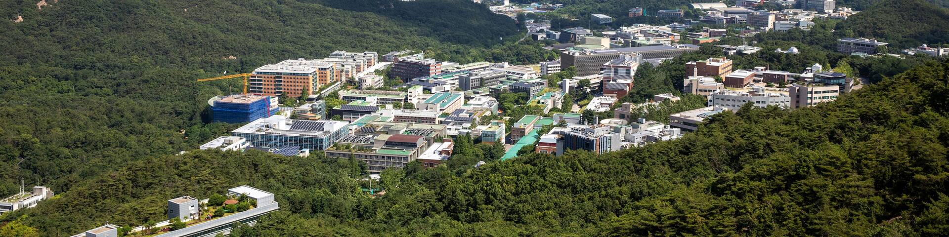 A cityscape with dense urban buildings, surrounded by green hills and a clear sky.