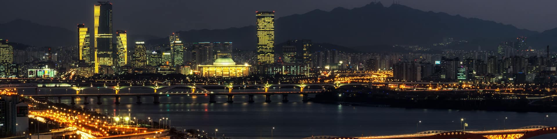 A nighttime cityscape with illuminated bridges and highway lights reflecting on the water.