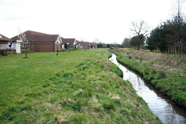 A narrow water channel running alongside a grassy area with houses in the background.