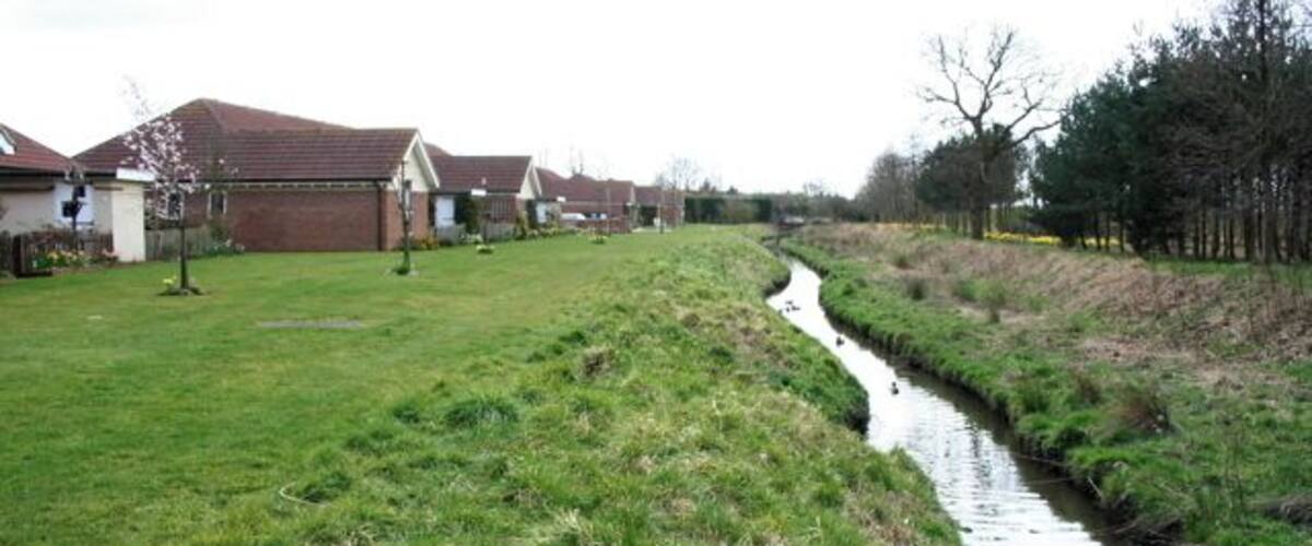 A narrow water channel running alongside a grassy area with houses in the background.