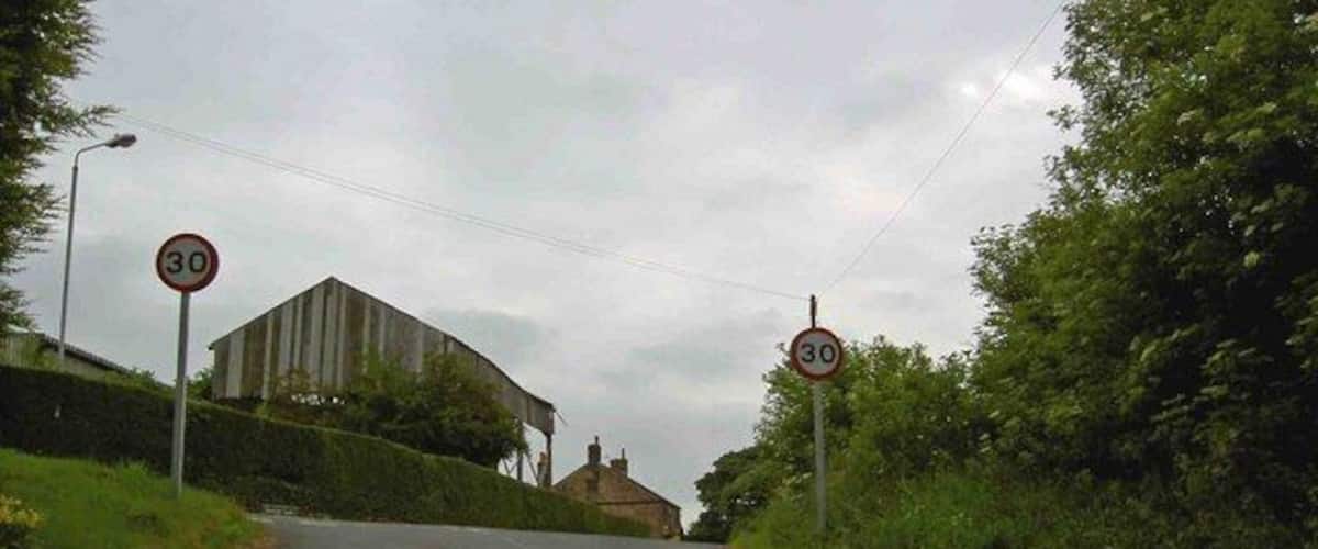 A rural road with a 30 mph speed limit sign on both sides, surrounded by greenery and a building in the distance.