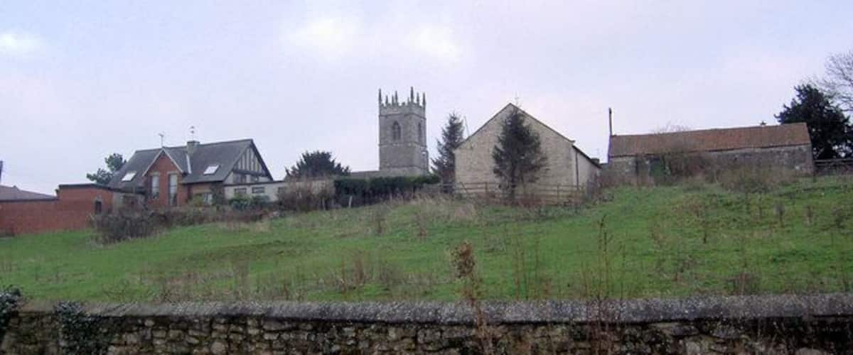 A rural landscape with a stone wall, a church with a tower, and a few residential buildings.