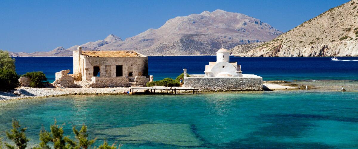 A coastal scene with a small white building and a pier extending into turquoise waters, surrounded by clear skies and distant mountains.