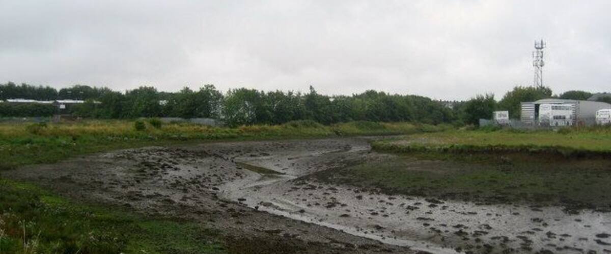 A muddy field with a water channel, a distant building, and a transmission tower.