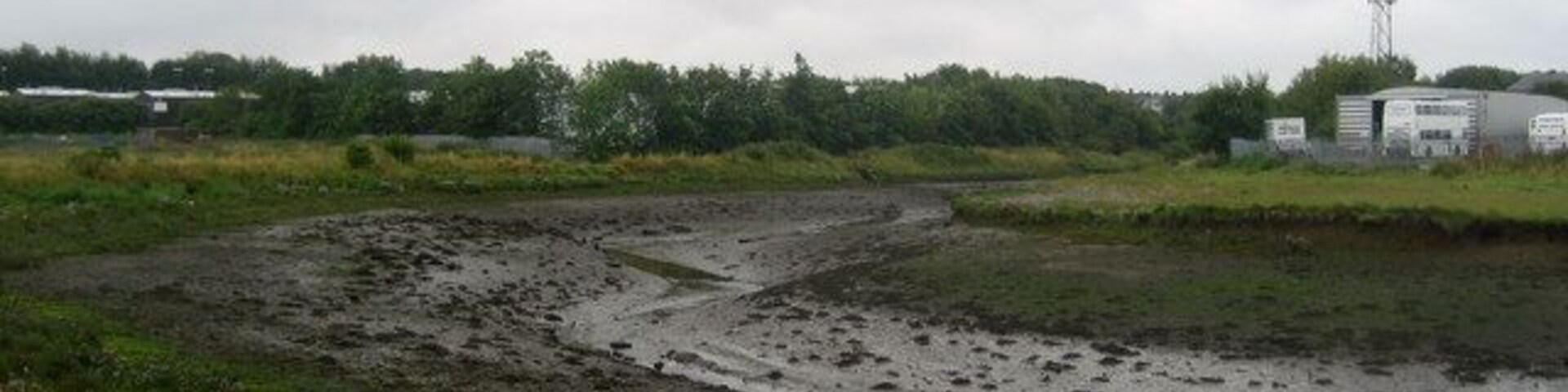 A muddy field with a water channel, a distant building, and a transmission tower.