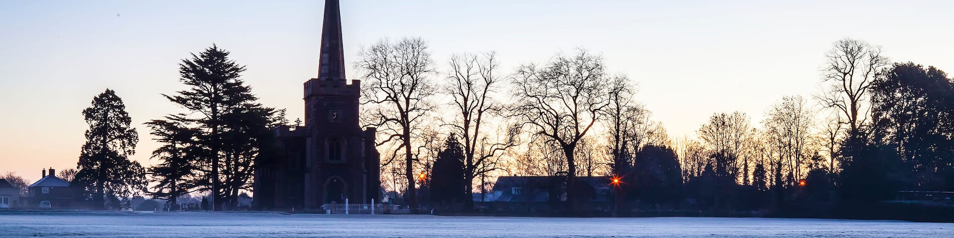 A church spire silhouetted against a sunset, a frost-covered field, and leafless trees.