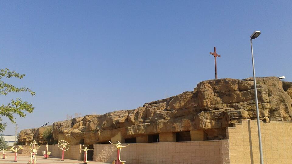 A rocky outcrop with a cross on top, outdoor exercise equipment, and a paved area.